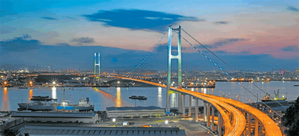 Ulsan Bridge illuminated at dusk over the harbor, with ships and city lights in the background